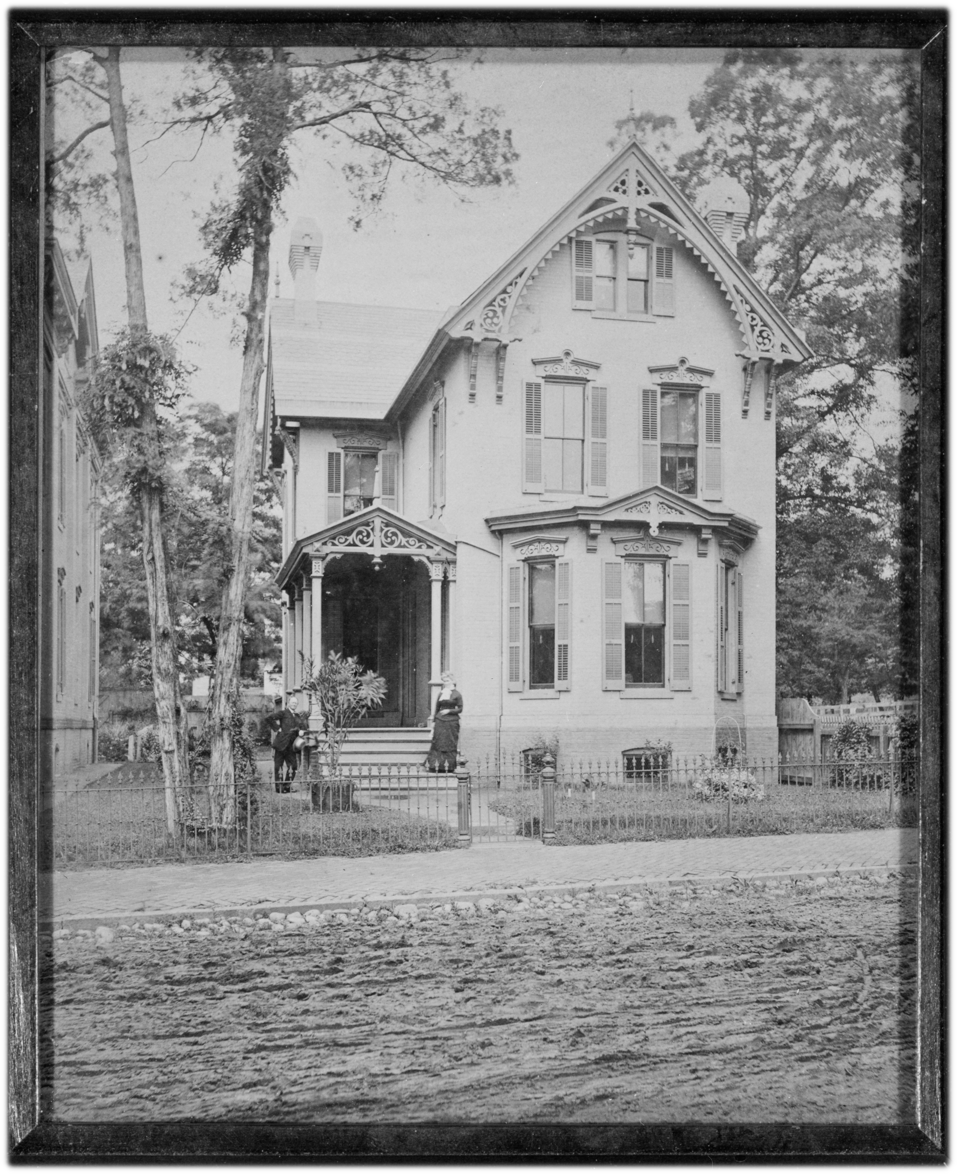 A black-and-white photograph of a classic three-story Victorian house, with a covered porch to the left, a bow window on the front, and ornate trim on porch and peaked roof line.