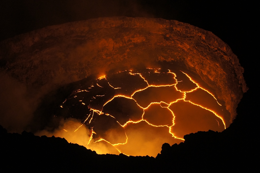 The floor of this crater on Kilauea is covered in a network of bright yellow-orange fissures. Steam rises from the bottom of the crater and the sides reflect an ominous orange glow from the fissures below.