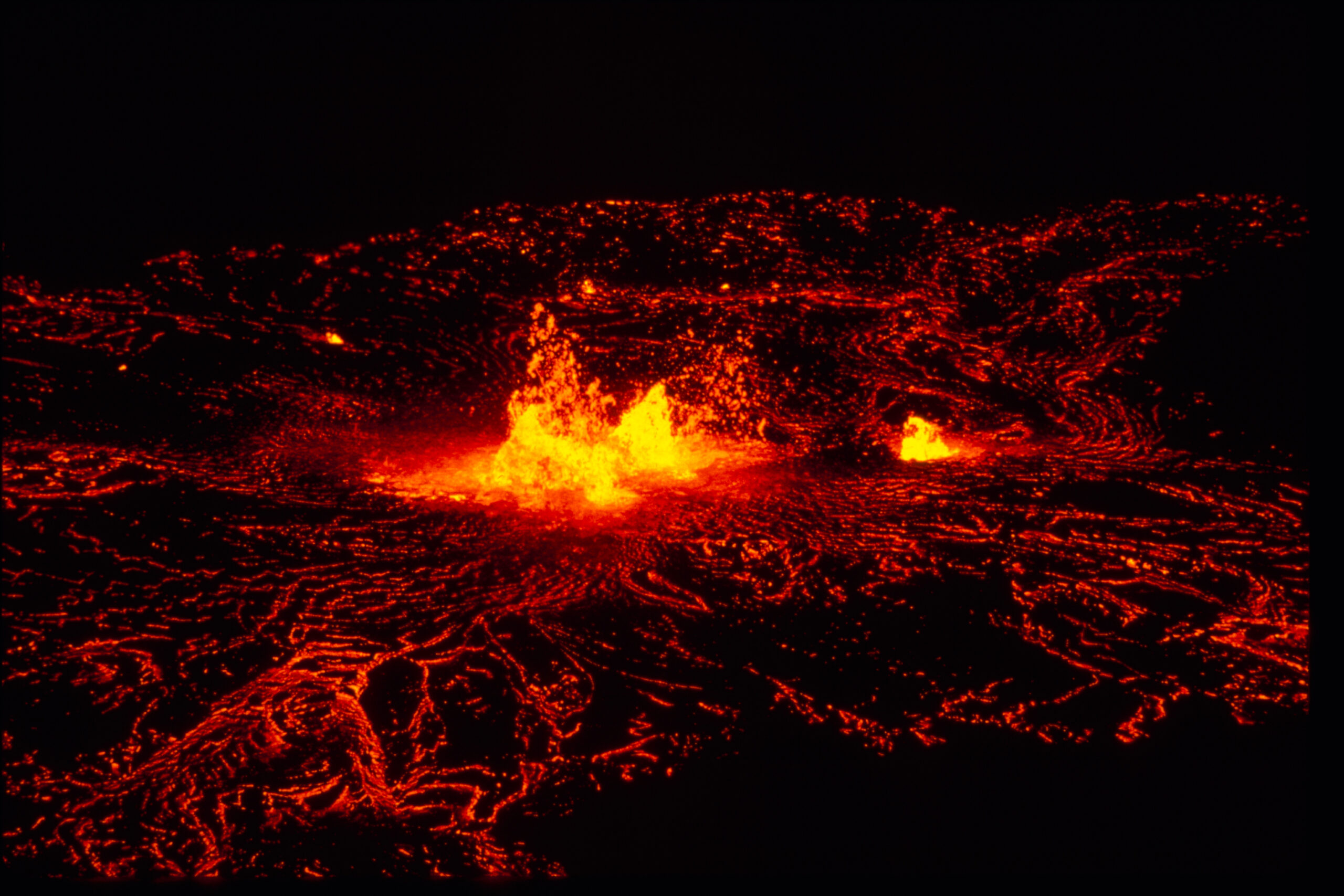 On a dark night, lava fountains up in three red-gold sprays.  A network of lacelike glowing lava strands surrounds the fountains on all sides, outlining the contours of the earth beneath. 
