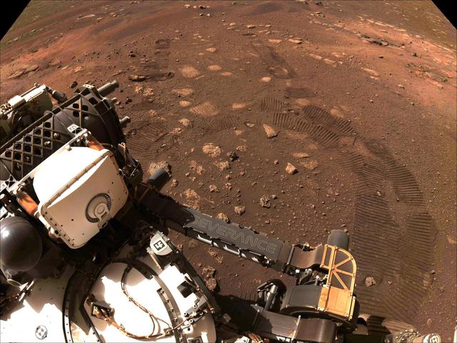 Part of the Perseverance rover gleams against the rocky red backdrop of Martian soil.   