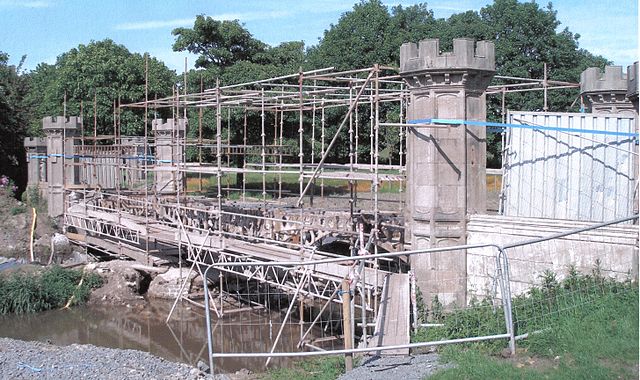 Four classic gray stone towers with battlements rise on either side of a piece of water.  Extensive scaffolding between the sides shows the bridge flooring in progress.  The blue sky and lush trees in the background add a pastoral note to the scene.