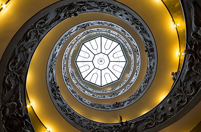 This upward-gazing photo of the spiral staircase in the Vatican museum emphasizes the dark carving of the staircase against the yellow sides. Light streams through a circular window marked in eight chevrons, forming the perfect focal point.