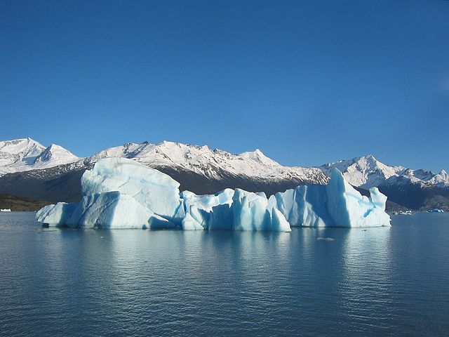 An irregular blue-white iceberg floats in the chilly waters off Argentina.  Snow-brushed mountains appear in the background. The lightly rippling ocean waters reflect the clear blue sky above in this bright image.