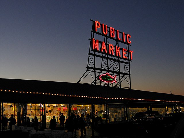 The words "Public Market" appear in vibrant, glowing red against a clear sunset sky.  The sign towers over a long, low-roofed building.  Many shoppers are silhouetted against the bright interior lighting.  Pike's Place Market, Seattle, runs late and colorfully.