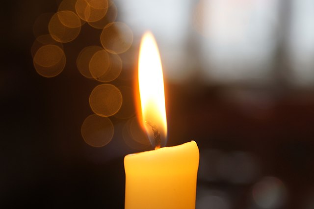 A close-up of a warm yellow candle flame. The creamy candle and bright flame are sharp against a half-dark background in this contemplative image.