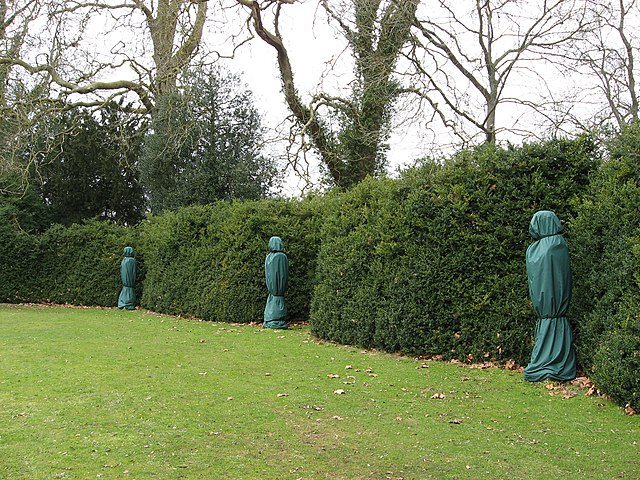 A large grassy lawn is surrounded by high evergreen hedges.  Statues, cloaked in green tarps, stand at intervals around the green.  Bare trees peep over the top of the hedge.  The mood is mysterious.